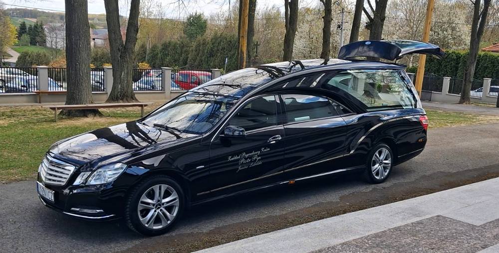 Czarny karawan Mercedes zakładu pogrzebowego Znicz przed kościołem, wśród drzew i krzyży